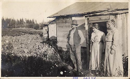Photo of the old Buzby farm, showing Harry and Louisa Buzby and daughter Marian standing outside a small farm building, with lush vegetables growing in the background.